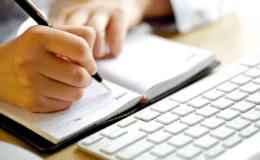 Close-up of a person writing in a planner next to a white keyboard.