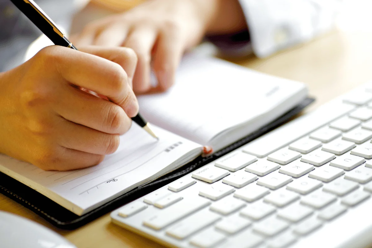Close-up of a person writing in a planner next to a white keyboard.