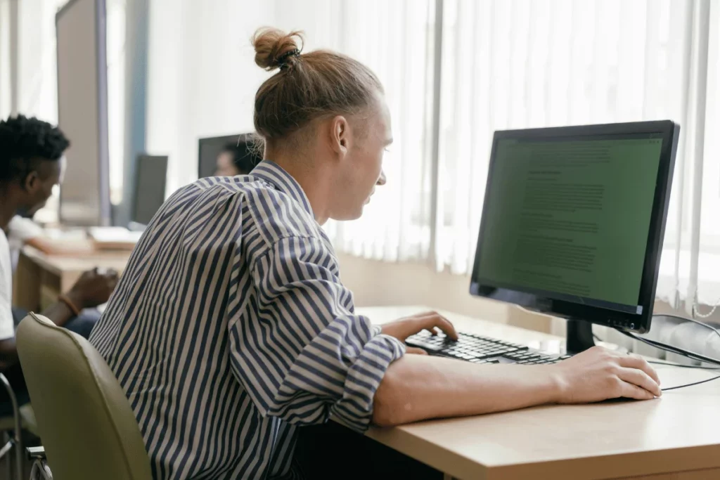 Man in striped shirt working on desktop computer in bright classroom setting.