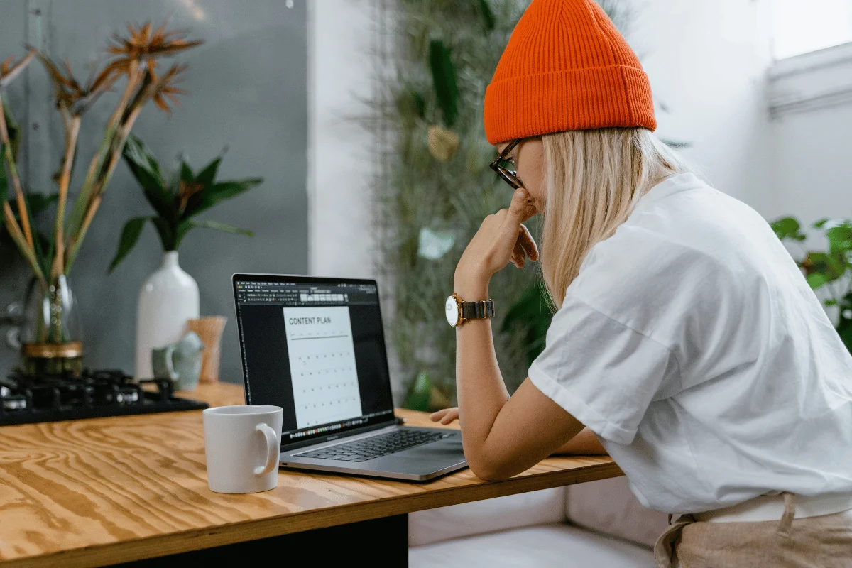 Woman in a beanie reviewing a content calendar on her laptop screen at a desk with a coffee mug.