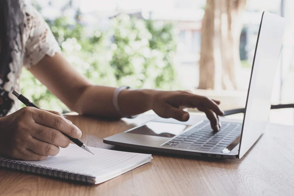 Individual multitasking by writing in a notebook and using a laptop.