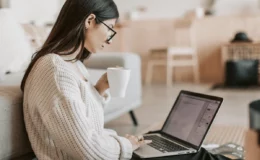 A woman with long dark hair and glasses sits on a couch, holding a coffee mug, focused on a laptop. The room has a cozy, relaxed atmosphere.