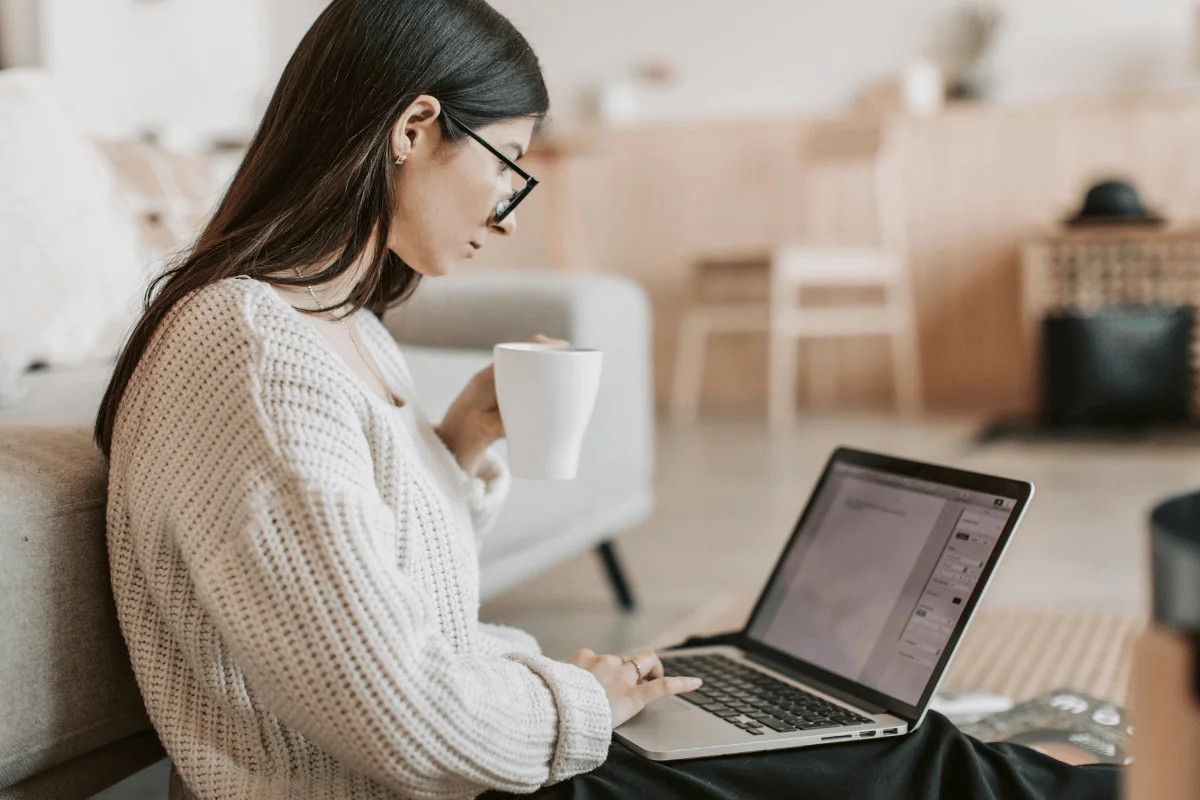 A woman with long dark hair and glasses sits on a couch, holding a coffee mug, focused on a laptop. The room has a cozy, relaxed atmosphere.