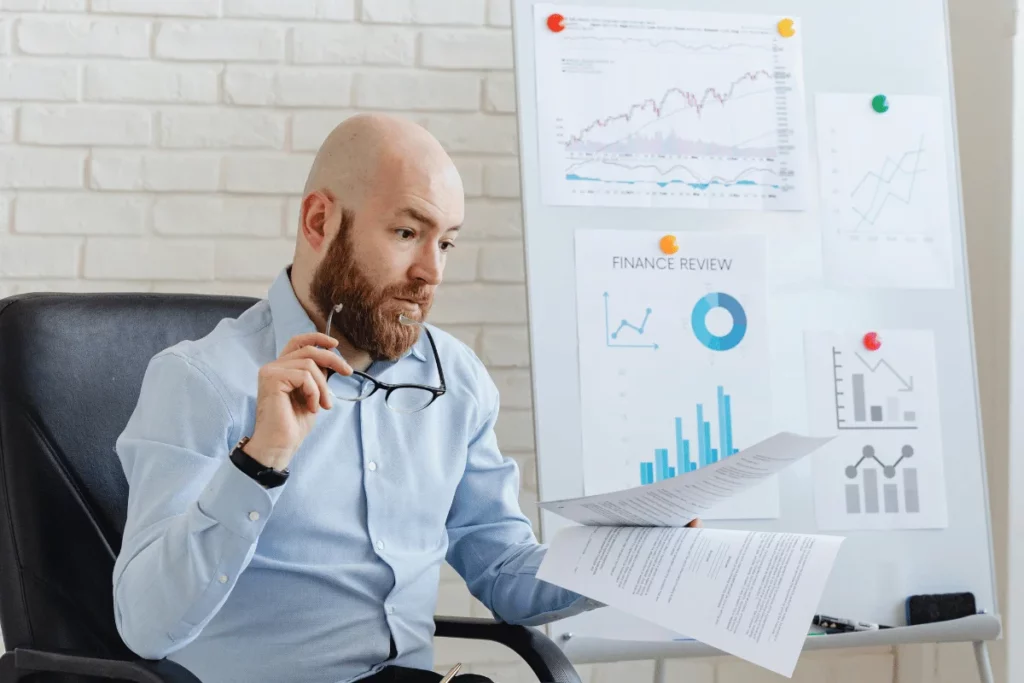 Hombre analizando informes impresos delante de un tablero de gráficos financieros.
