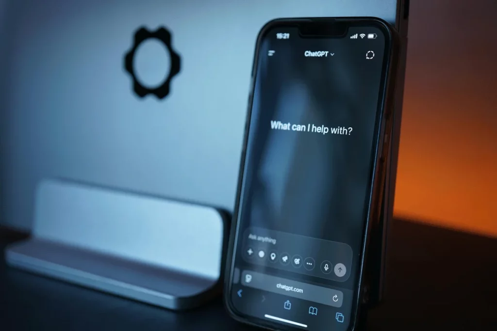 A smartphone displaying the ChatGPT interface, showing the prompt "What can I help with?" on a dimly lit desk.