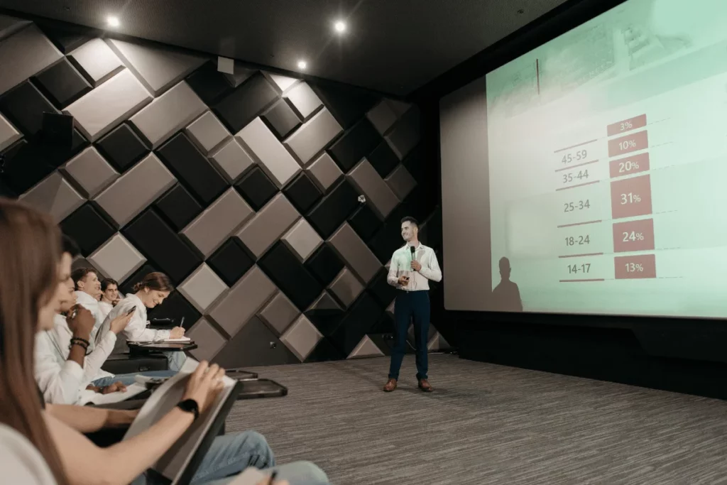 Presenter speaking in front of an audience with a slide displaying demographic data on a large screen.