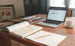 A cozy workspace with a wooden table, open laptop, sketchbook, pencil, and coffee cup.