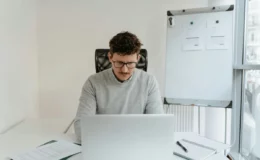Office worker using computer with paperwork and whiteboard in view.