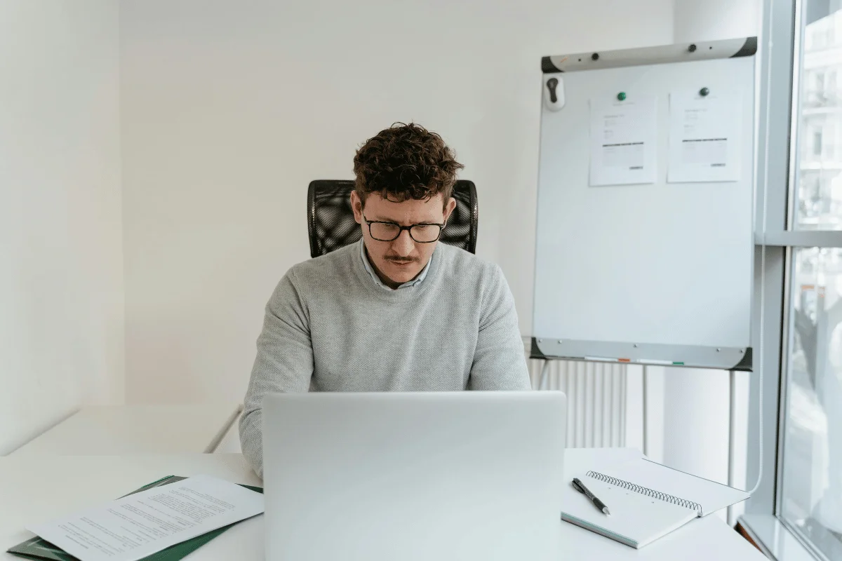 Office worker using computer with paperwork and whiteboard in view.